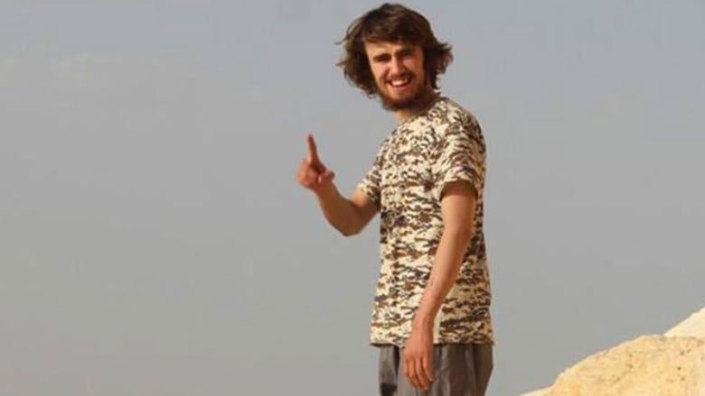 Jack Letts in Syria with a beard and long brown hair looking toward the camera, wearing cargo pants and a camouflaged shirt, pointing his index finger toward the camera.