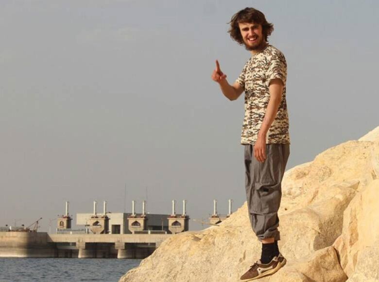 Jack Letts in Syria with a beard and long brown hair looking toward the camera, wearing cargo pants and a camouflaged shirt, pointing his index finger toward the camera.