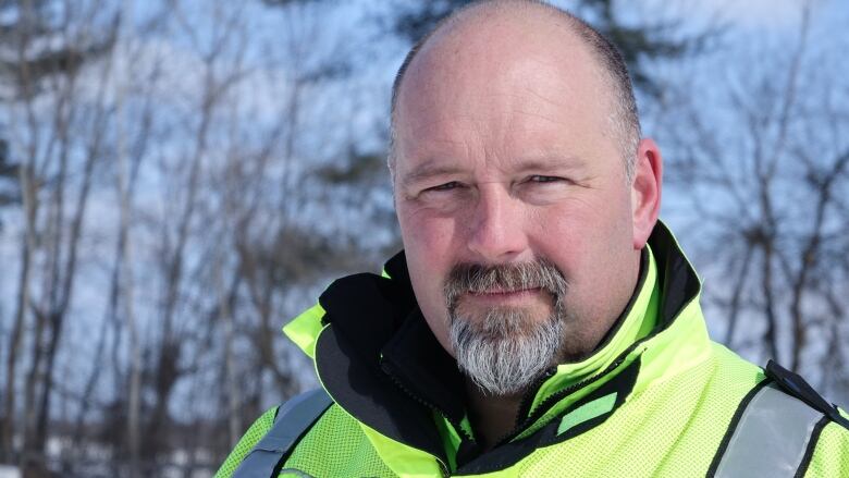 A close-up of a middle-aged man wearing a bright yellow jacket, outdoors in winter, in front of a grove of trees.