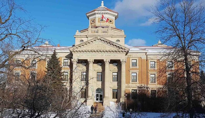 Large neoclassical style orange building with white panneling. A canadian flag hangs from the builing and the building is shrouded by trees. It's winter, and there is snow on the ground and the trees don't have leaves.