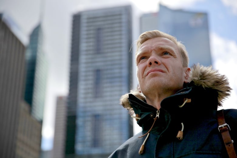 A man wearing a winter jacket stands in front of large buildings.