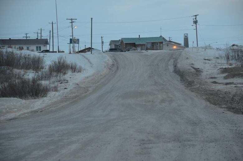 A frozen dirt road goes up a hill past some snowbanks towards some buildings on the horizon. 