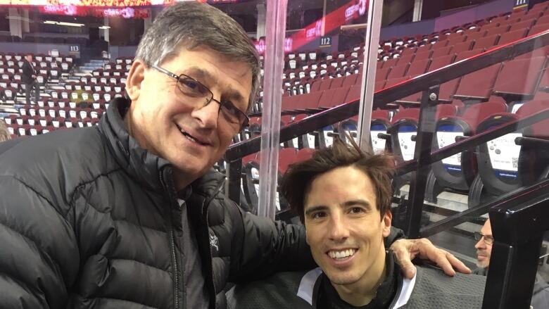 An older man with glasses puts his arm around a younger hockey player wearing goalie equipment.