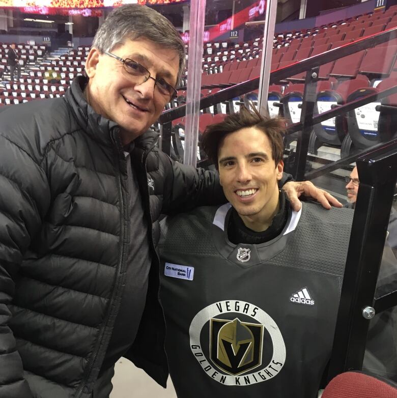 An older man with glasses puts his arm around a younger hockey player wearing goalie equipment.