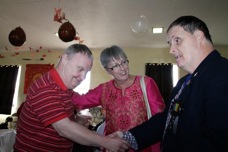 Two men with Down syndrome shake hands while a woman looks on during a celebration. 