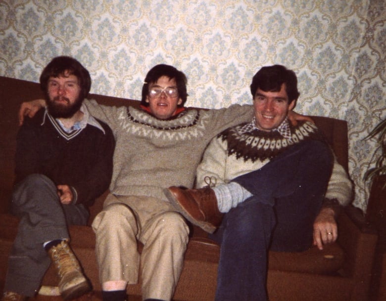 Three men sit on a sofa, arms around each other's shoulders, in a photo from the early 1980s. 