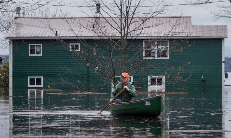 A man in an orange hat paddles a green canoe in floodwaters in front of a green flooded home.