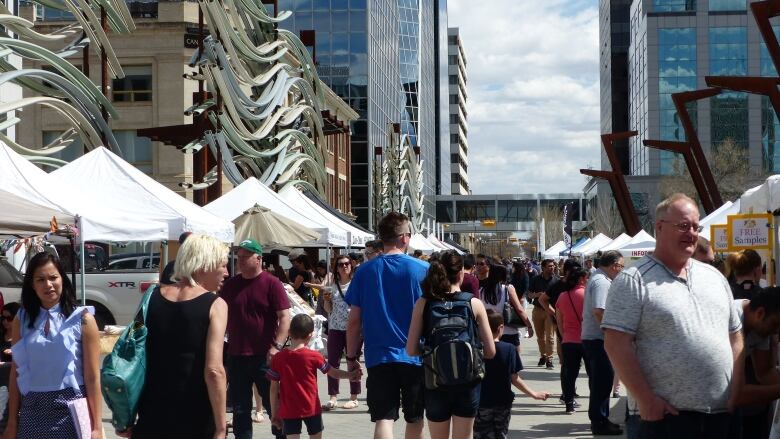 Regina Farmers' Market goers fill a plaza as vendors are set up on each side with skyscrapers towering overtop. 