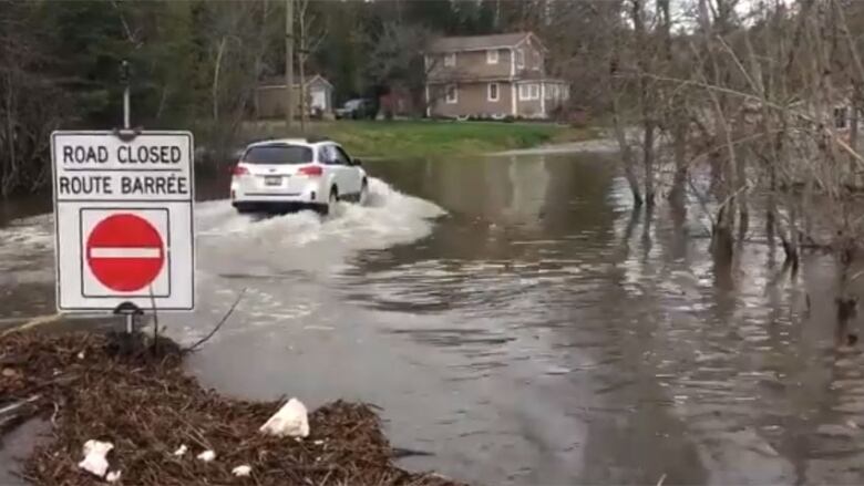 A white SUB drives through high floodwaters in front of a sign that says the road is closed.  