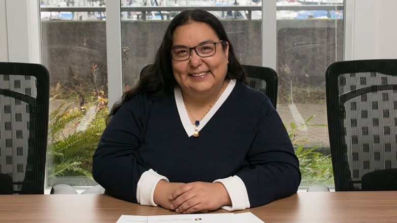 A smiling Indigenous woman with glasses and long dark hair sits at a desk with her hands clasped in front of her.