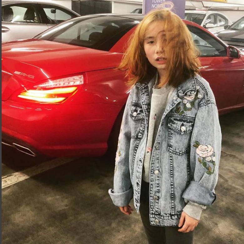 A young girl with shoulder-length blonde hair stands in a denim jacket in front of a red luxury car in a parking garage.