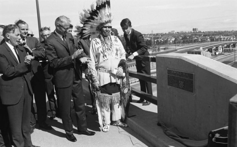 Men in suits and a man in Indigenous chief regalia look at a highway plaque.