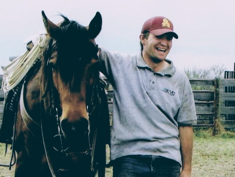 A young man smiles and looks relaxed with his arm around a horse, wearing a t-shirt, jeans and baseball cap.