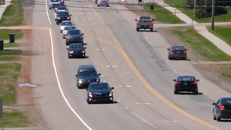 An aerial shot shows a funeral procession led by a hearse. 