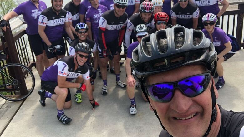 A man in a bike helmet poses for a selfie with a dozen or so other people in helmets and bike suits.