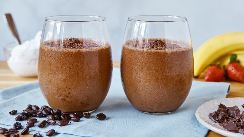 2 clear glasses with chocolate smoothies in them, on a light blue napkin in front of a light blue background. Bananas and strawberries are in the background, and coffee beans are in the foreground.  