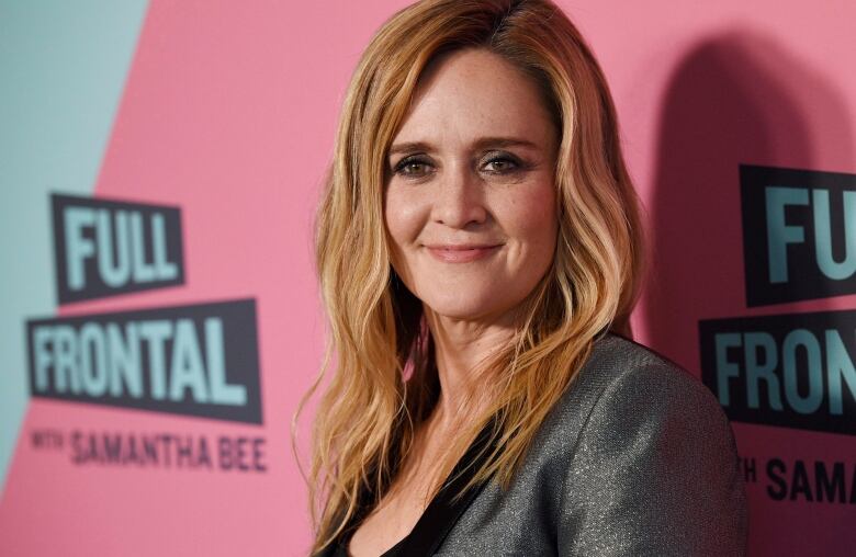 A woman with blond hair wearing a grey blazer stands before a pink backdrop.