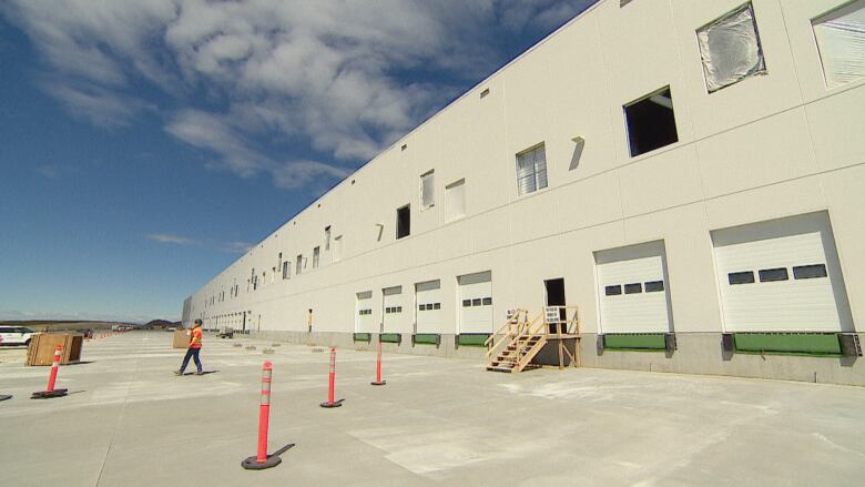 a large warehouse against a blue sky. a worker in a high visibility safety vest walks holding a box.