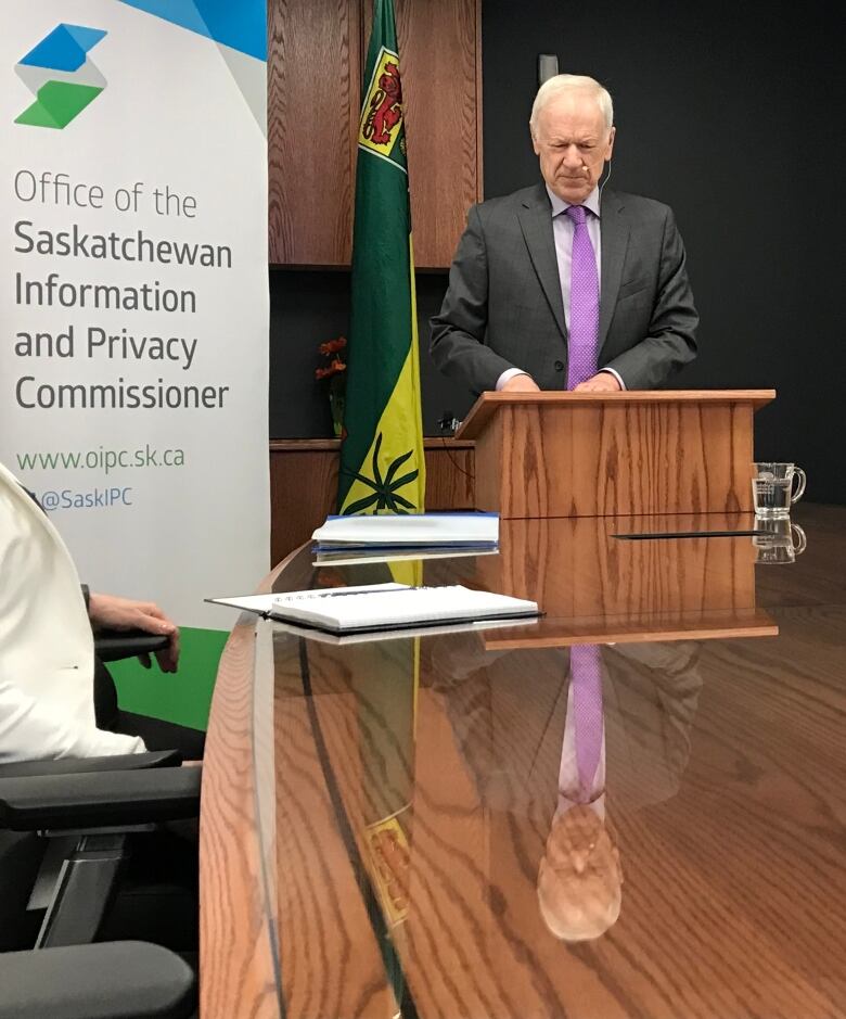 a man stands at a podium at the end of a table with a Saskatchewan flag behind him