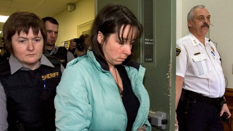 A woman with shoulder length brown hair and wearing a teal colored coat walks out of a room surrounded by court sheriffs and holding her head down.