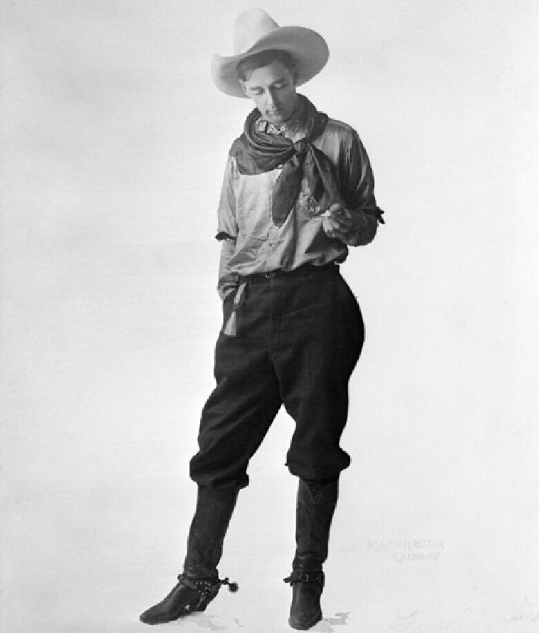 A tall, thin man dressed in cowboy boots and hat poses for a studio photograph. He appears to be looking down at an object in his left hand.