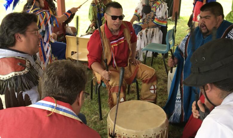 Five men sit around a drum in Indigenous regalia.