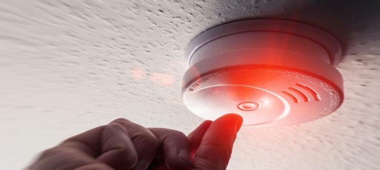 A smoke detector on a ceiling with the red light flashing and a hand reaching up towards it. 