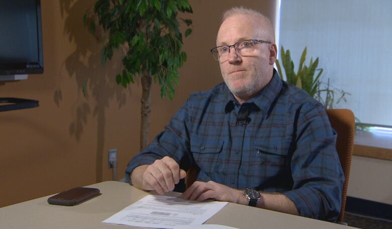 A guy with glasses sits at a table with a piece of paper and cell phone on the table