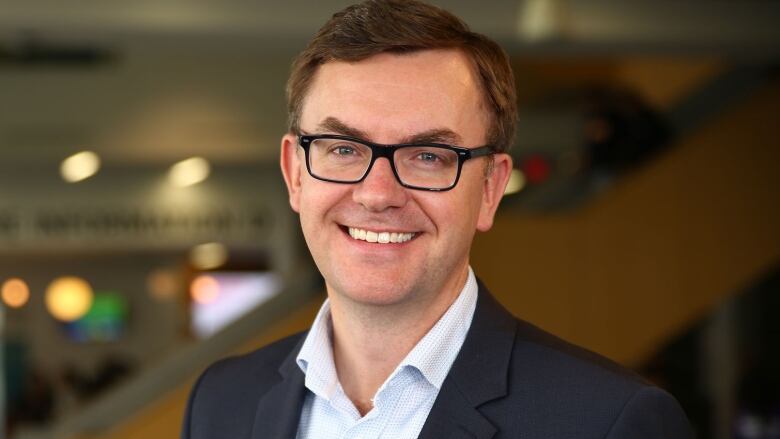 A man in a suit and glasses stands indoors