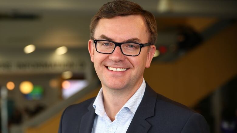 A man in a suit and glasses stands indoors