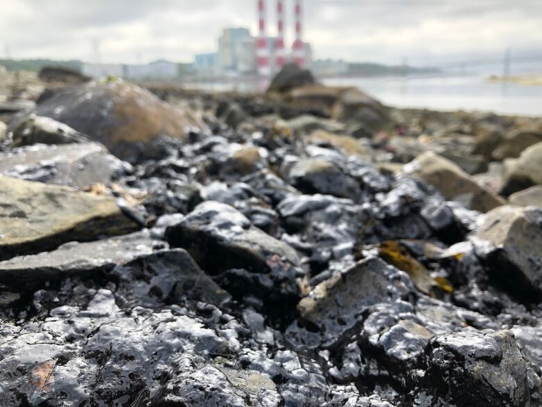 A rocky beach with water and smoke stacks in the background.