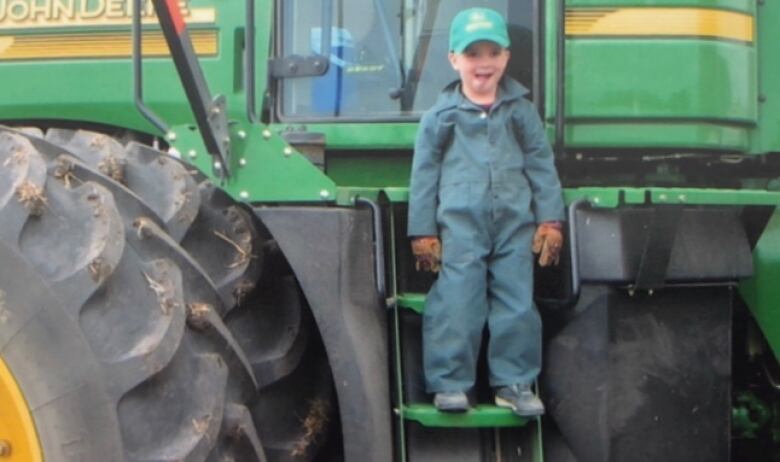 A little boy in blue coveralls stands on the steps of a green farm vehicle.