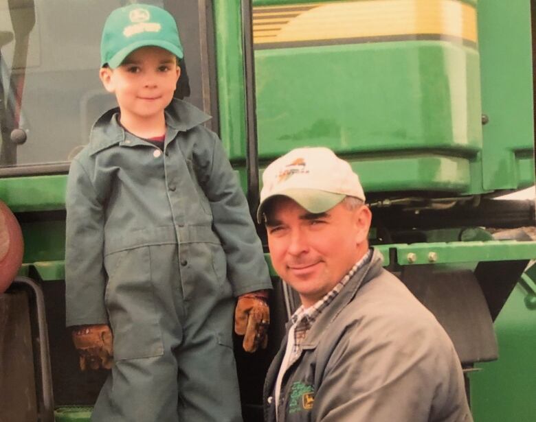 A young boy in coveralls stands on a farm vehicle, standing alongside a man kneeling.