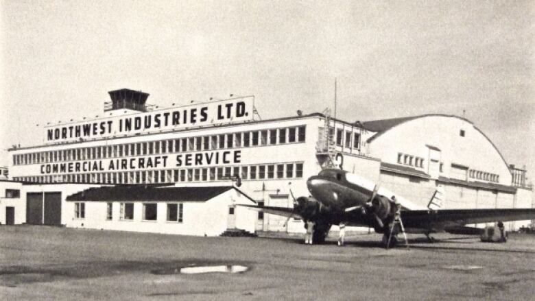 A black and white photo of an old airplane hanger with an airplane outside.