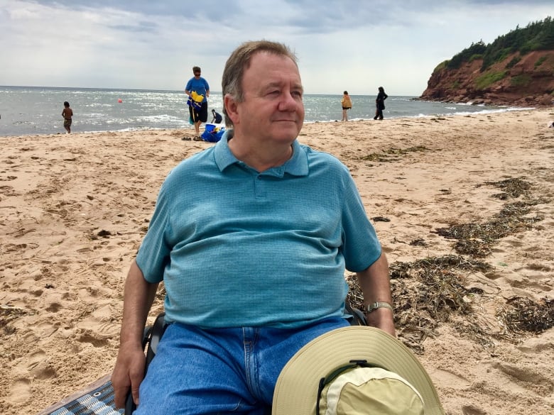 A man in a wheelchair sitting on a beach with people in the background. He is looking off-camera. 