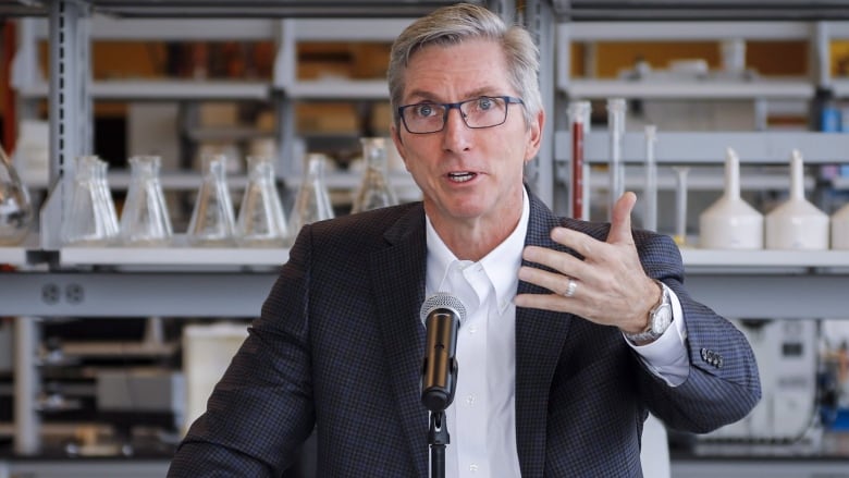 A close-up shot of a man with blonde hair and glasses speaking into a microphone.