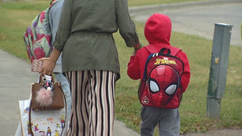 A woman walks hand in hand with a small child in a red jacket wearing a spiderman backpack.