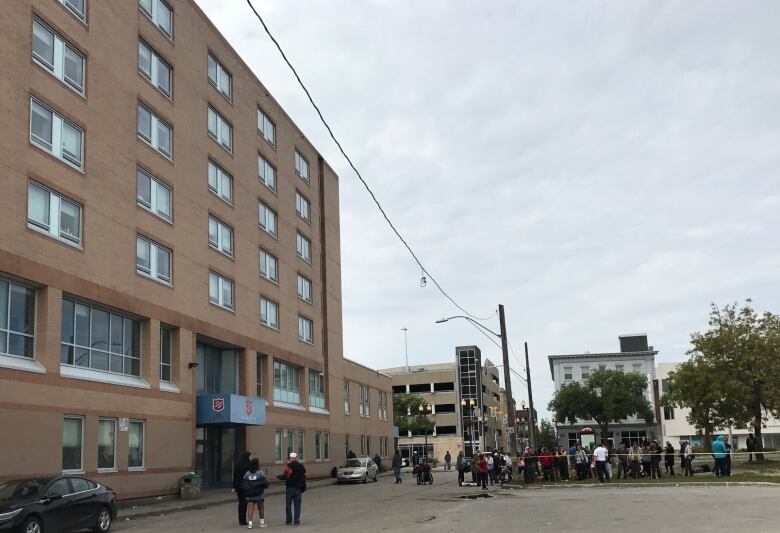 Photo shows a large beige building with a few people outside of it talking to each other. 