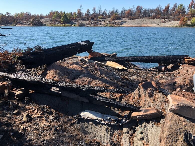 Some burnt remains of a house sit on a rocky island, with a river, rocks and trees in the background