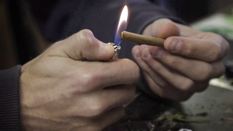 Fingers hold the end of a joint of marijuana under a flame.