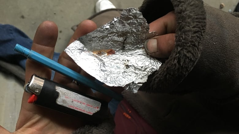 A person holds up a piece of aluminum foil with a brown substance on it, along with a lighter and a small pipe.