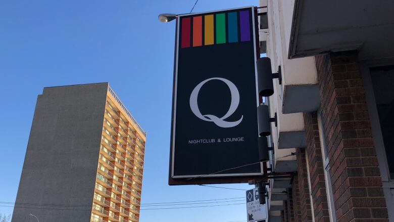 A black sign topped with vertical stripes in rainbow colours and featuring a large white letter Q is attached to the side of a brick building on an urban street.