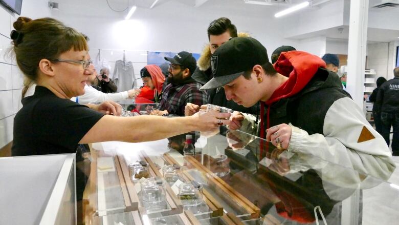 A man in a black cap leans over and sniffs a jar of cannabis, held in front of him by a woman behind the counter of a retail store. 