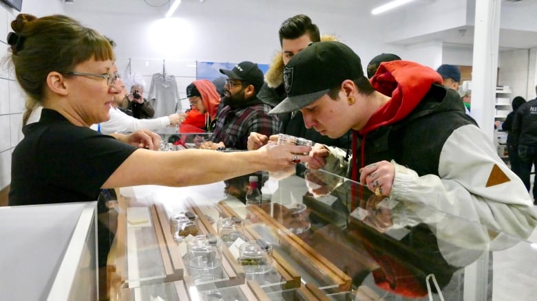 A man in a black cap leans over and sniffs a jar of cannabis, held in front of him by a woman behind the counter of a retail store. 