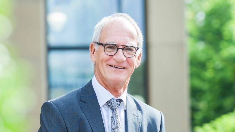 A white haired man with glasses wear a suit and stands outdoors, smiling at the camera.