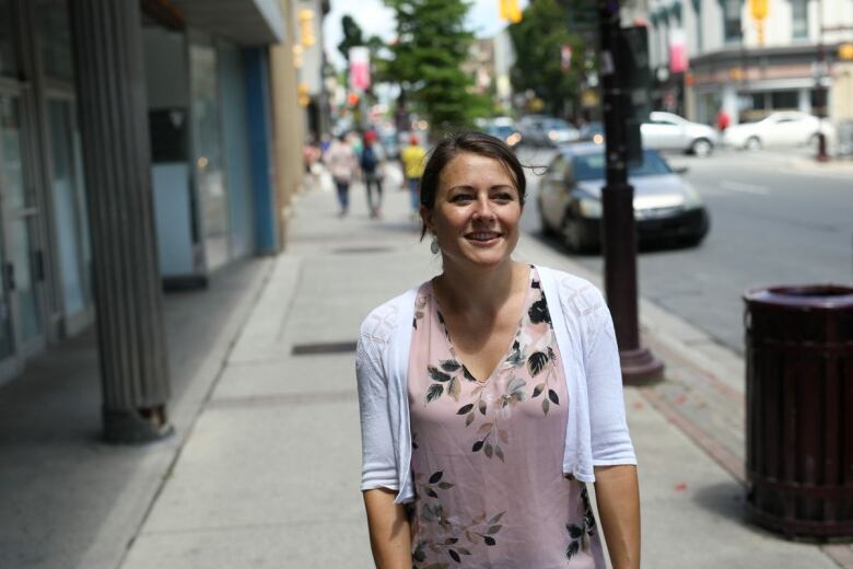 A woman walks on a city sidewalk wearing a floral top and white cardigan.