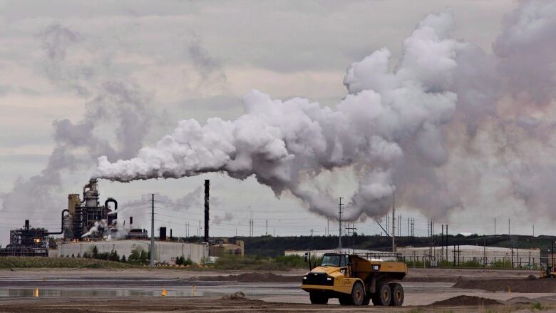 A dump trump stops in front of a large oil sands extraction facility. A large plume of smoke billows rightwarsd from a column.