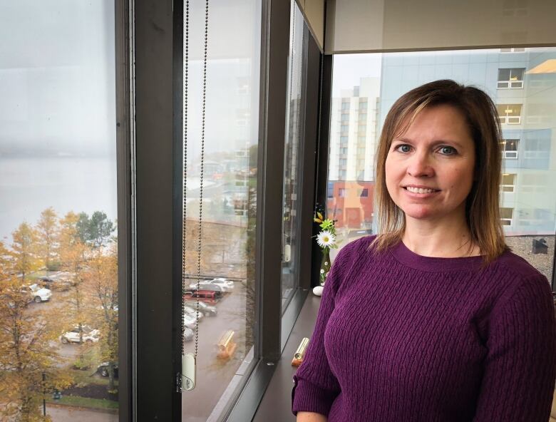 A woman with a purple sweater stands next to a window overlooking a harbour.