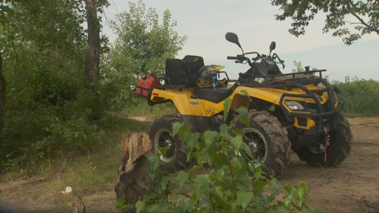 A yellow four-wheeler is parked on a dirt trail and surrounded by greenery.