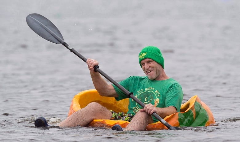 A man wearing a green toque and green shirt sits in a giant pumpkin that is floating on a lake. He holds a paddle.